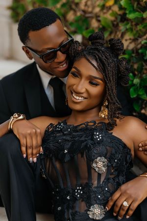 Smiling Black couple in elegant black outfits pose intimately outdoors, woman in lace dress with braided updo.