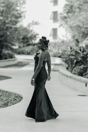 Black and white photo of a woman in an elegant mermaid gown with floral hair accessory posing outdoors.