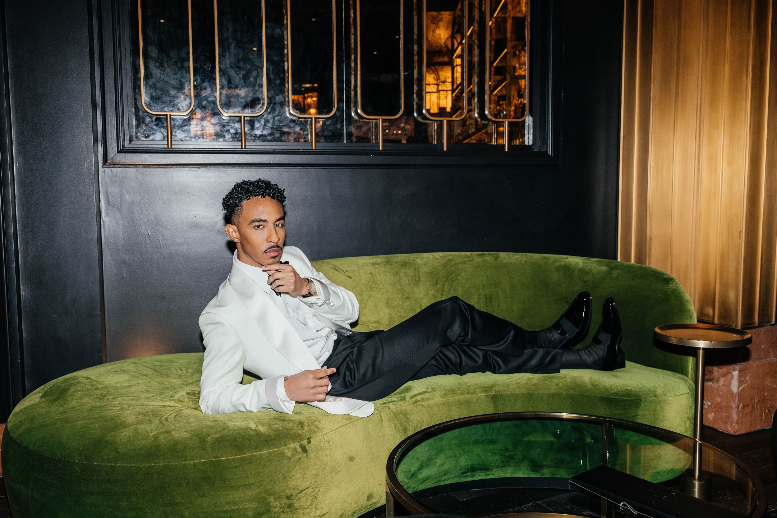Stylish man in white shirt and black pants relaxing on a green velvet sofa in a luxury interior.