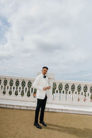 Dapper groom in white tuxedo jacket and black bow tie stands confidently on an outdoor terrace under cloudy skies.