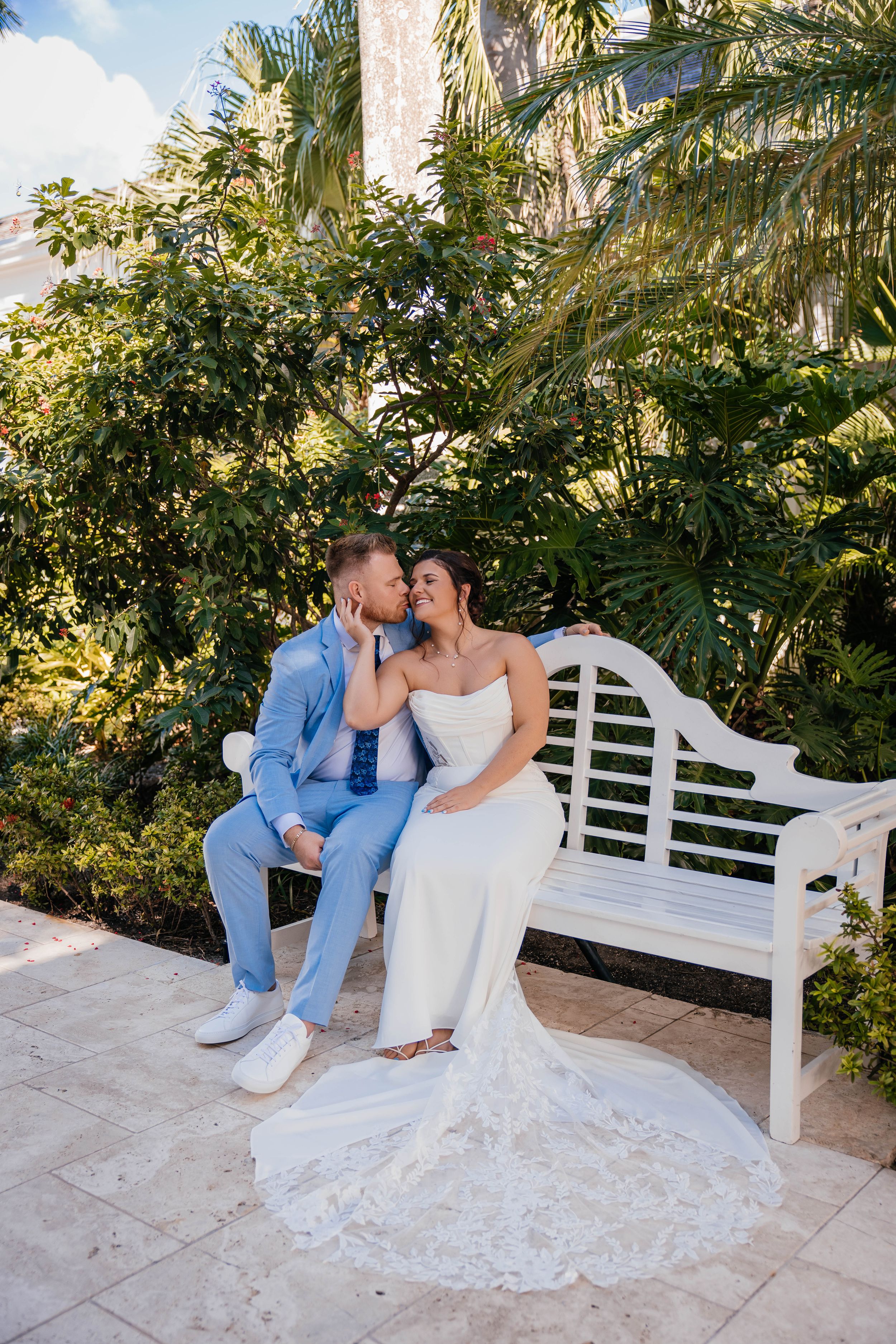 Bride and groom sharing a kiss on a white garden bench surrounded by tropical greenery.