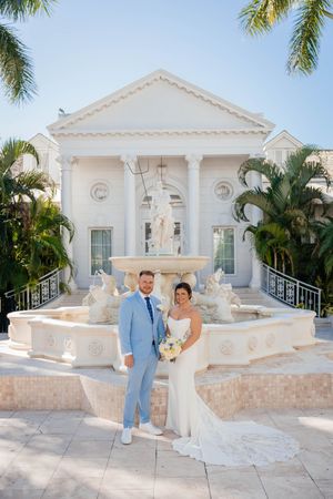 Bride and groom posing in front of a white neoclassical fountain with palm trees at a tropical wedding venue.