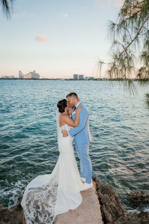 Bride and groom share a romantic kiss on rocky shoreline at sunset with turquoise ocean and city skyline behind them.