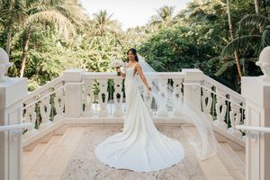 Bride in elegant white gown with long veil stands on ornate balcony surrounded by lush tropical greenery.