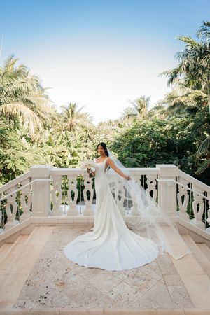 Bride in elegant white gown with long train poses on marble balcony surrounded by tropical palms on her wedding day.