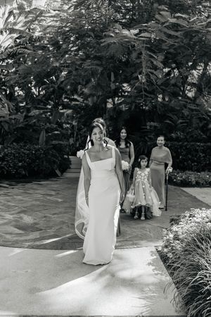 Black and white photo of a bride in a sleek white gown walking down a garden aisle with her bridal party.