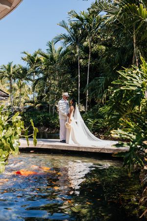 Bride and groom share a romantic moment on a koi pond dock surrounded by lush tropical palms at an outdoor wedding.