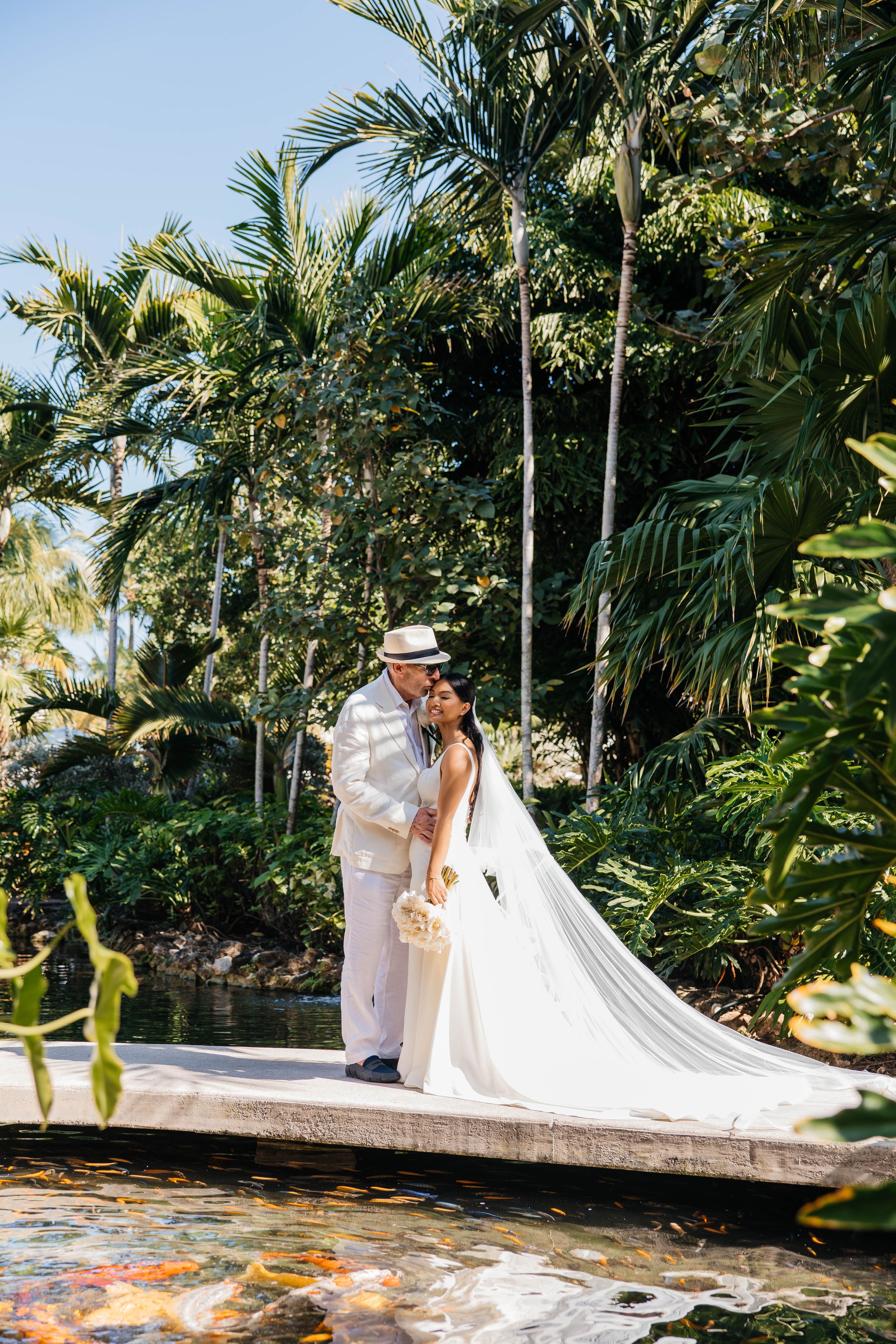 Bride and groom share a kiss on a wooden dock surrounded by a koi pond and lush tropical palm trees.