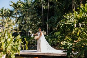 Bride in a flowing white gown stands on a wooden deck surrounded by lush tropical palm trees and greenery.