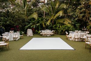 Outdoor tropical wedding reception with white dance floor, round tables, gold chairs, and lush greenery backdrop.