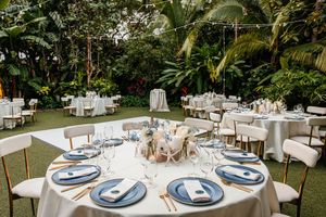 Elegant outdoor wedding reception with round tables, blue charger plates, and lush tropical garden backdrop.