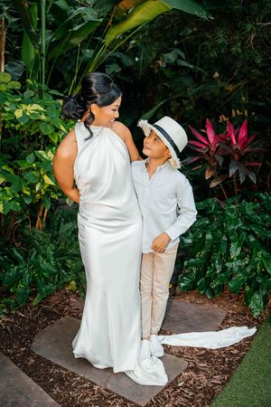Bride in white halter gown poses with young boy in hat and light suit amid lush tropical greenery.