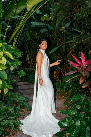 Bride in elegant white satin halter gown holding a wine glass, posing among lush tropical greenery.