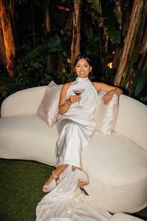 Elegant woman in white satin gown holding wine glass, seated on cream curved sofa amid lush tropical greenery.