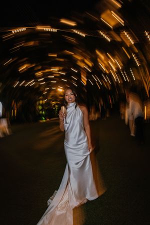 Bride in silver gown stands in illuminated tunnel with swirling bokeh lights at night.