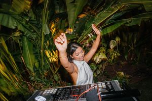 Female DJ performing outdoors surrounded by tropical plants, arms raised joyfully behind a mixer at a garden event.