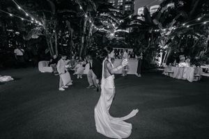 Bride in white gown dancing at elegant outdoor wedding reception with string lights and tropical trees in black and white.