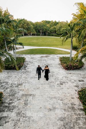 Aerial view of couple in black formal wear holding hands walking up stone pathway flanked by palm trees.