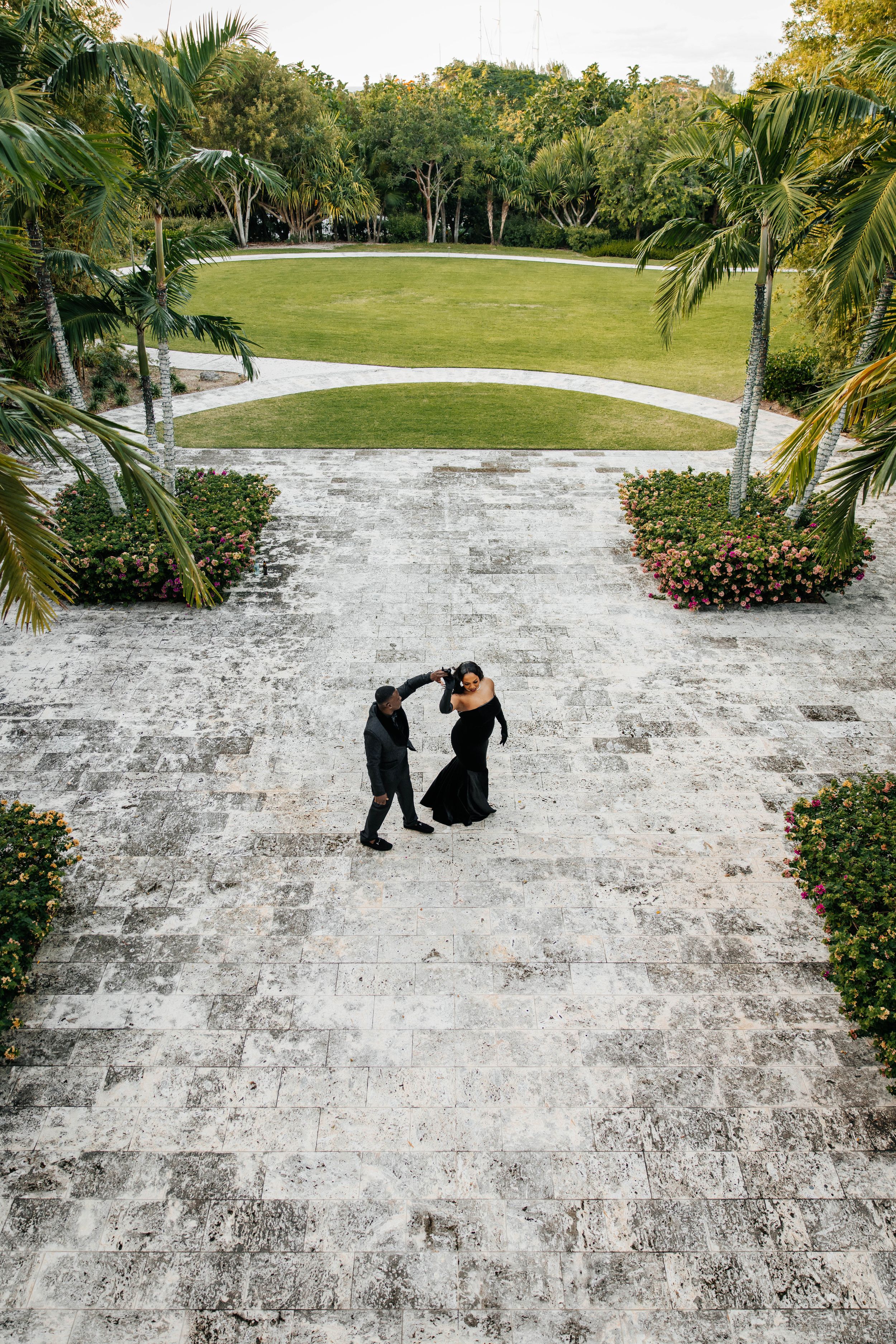 Aerial view of a couple dancing in black formal attire on a stone plaza surrounded by tropical gardens.