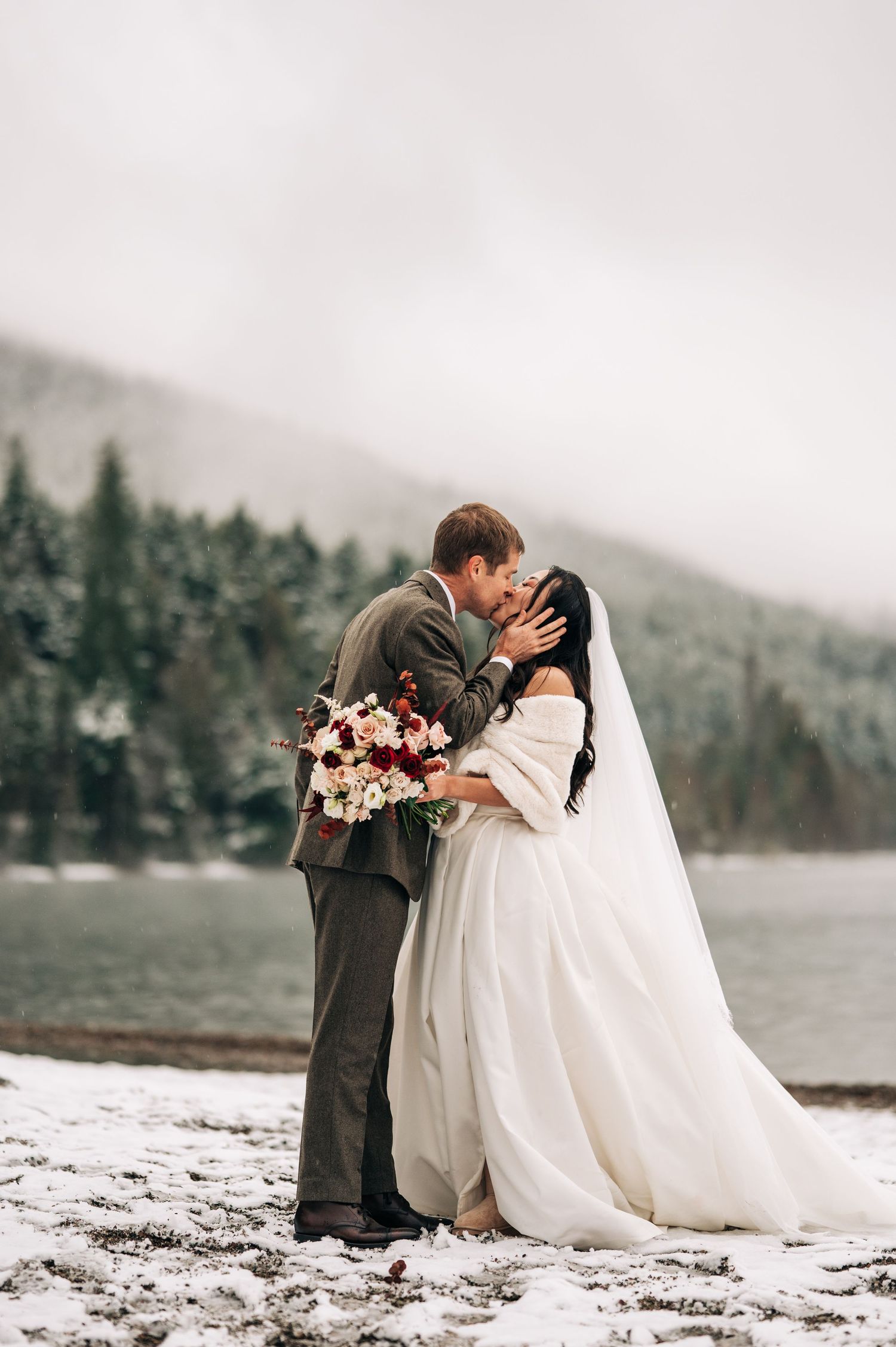 Bride and groom share a kiss in a snowy winter landscape, bride holding a red and white bouquet with pine trees behind.