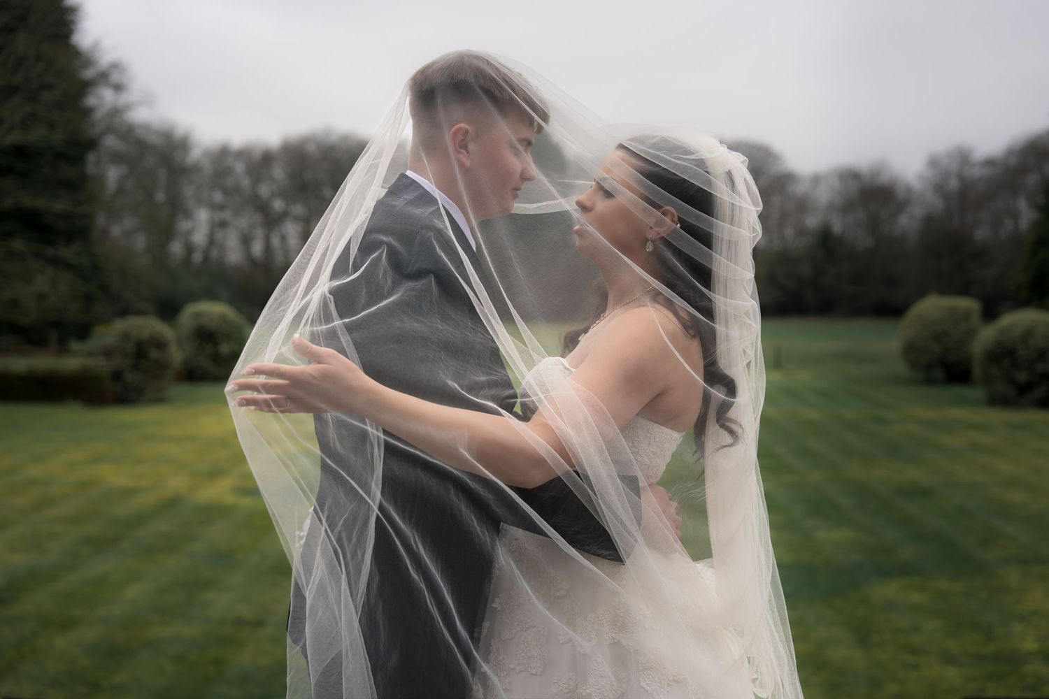 Bride and groom share romantic moment under flowing wedding veil outdoors in lush green garden setting.