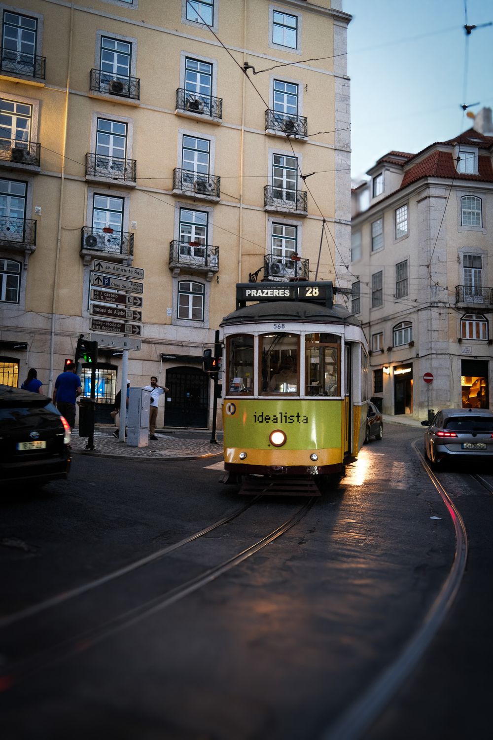 Historic yellow tram 28 to Prazeres navigating narrow Lisbon streets at dusk, passing classic Portuguese architecture.