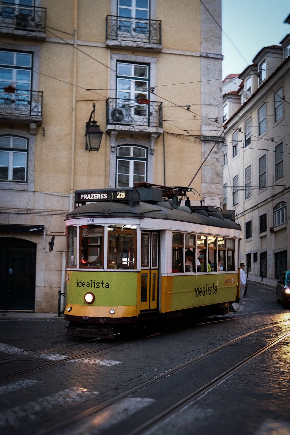 Yellow tram 28 to Prazeres navigating narrow Lisbon streets past historic buildings at dusk.