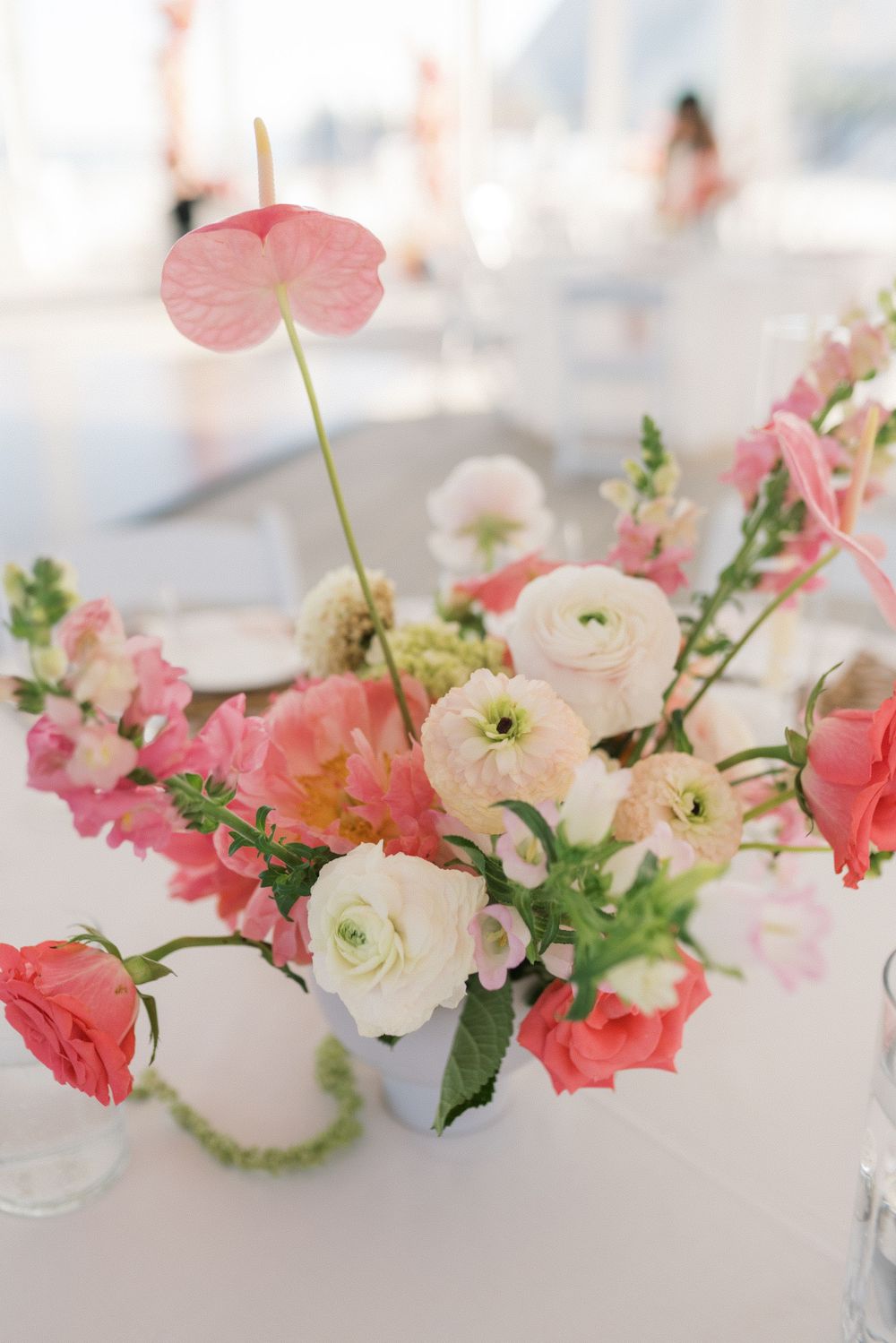 Pink and white floral centerpiece with ranunculus, anthurium, and roses in a white vase on a wedding reception table.