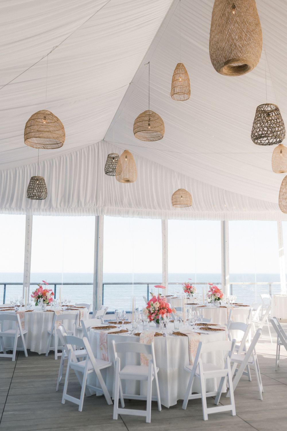 Elegant beachside wedding reception tent with wicker pendant lights, white chairs, and coral floral centerpieces overlooking the ocean.