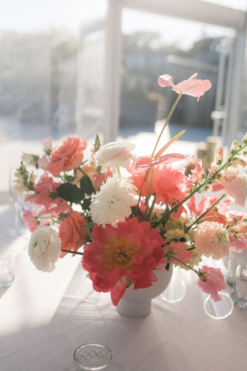 Elegant coral and white floral centerpiece in white vase on white tablecloth, bathed in natural sunlight.