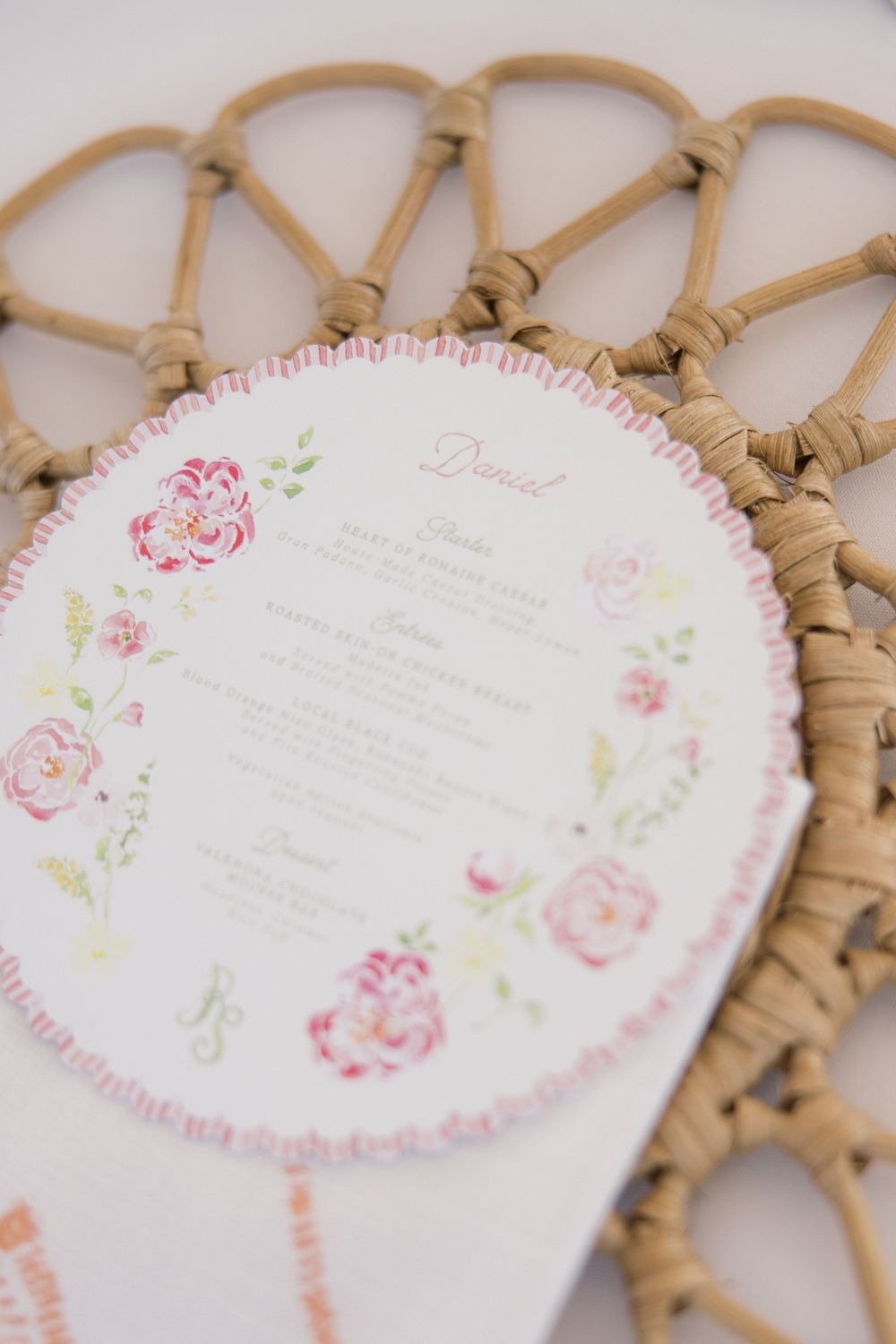 Circular floral wedding menu card with pink roses on woven rattan charger plate.