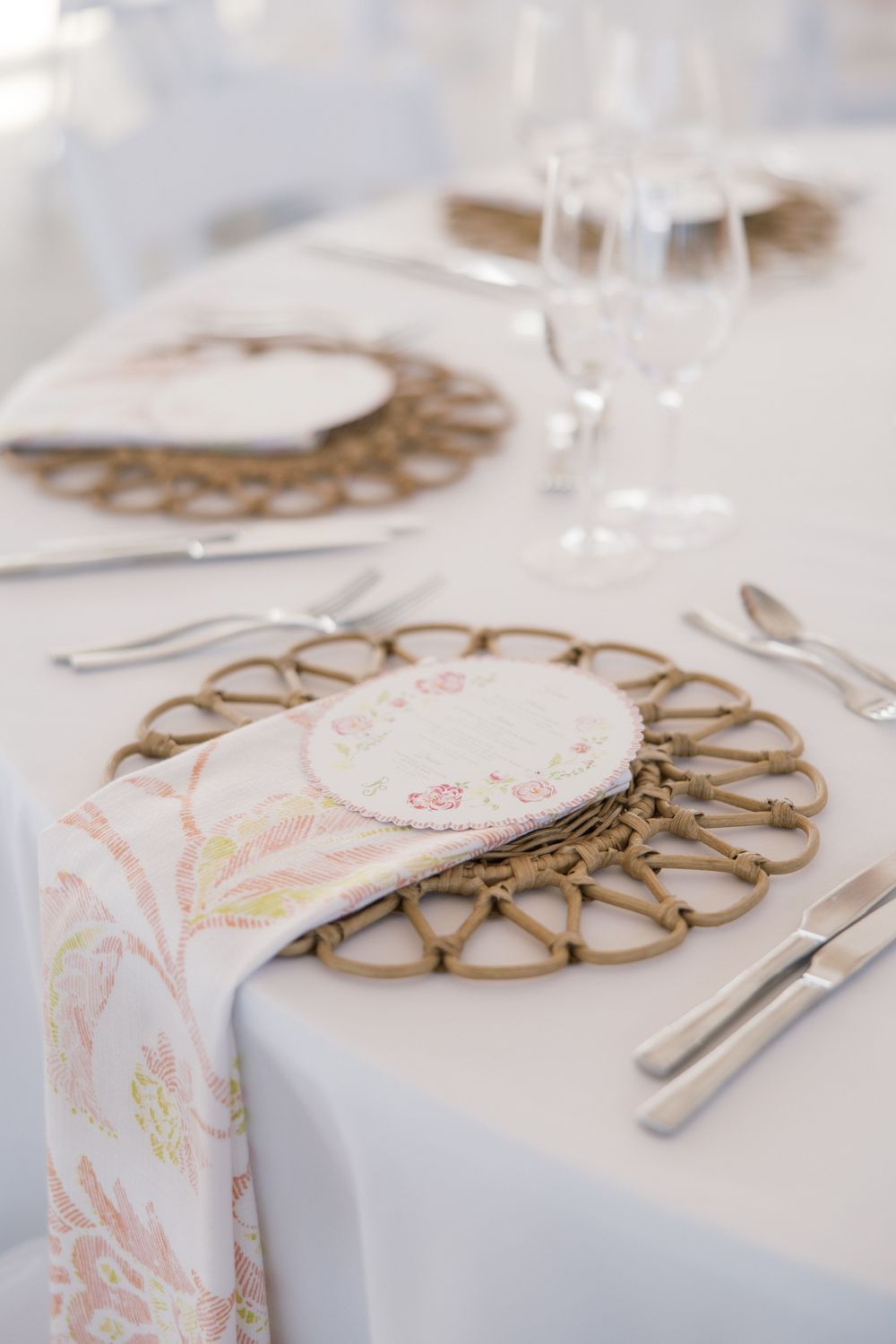 Elegant table setting with woven rattan charger plates, floral napkins, and crystal glasses on a white tablecloth.
