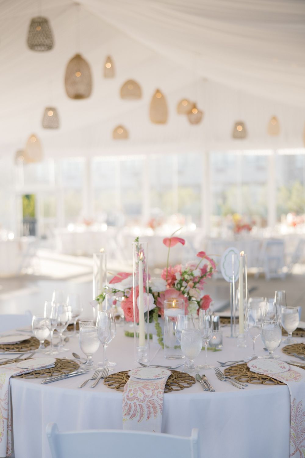 Elegant wedding reception table with pink floral centerpiece, white candles, gold chargers, and hanging rattan lanterns in a tent.