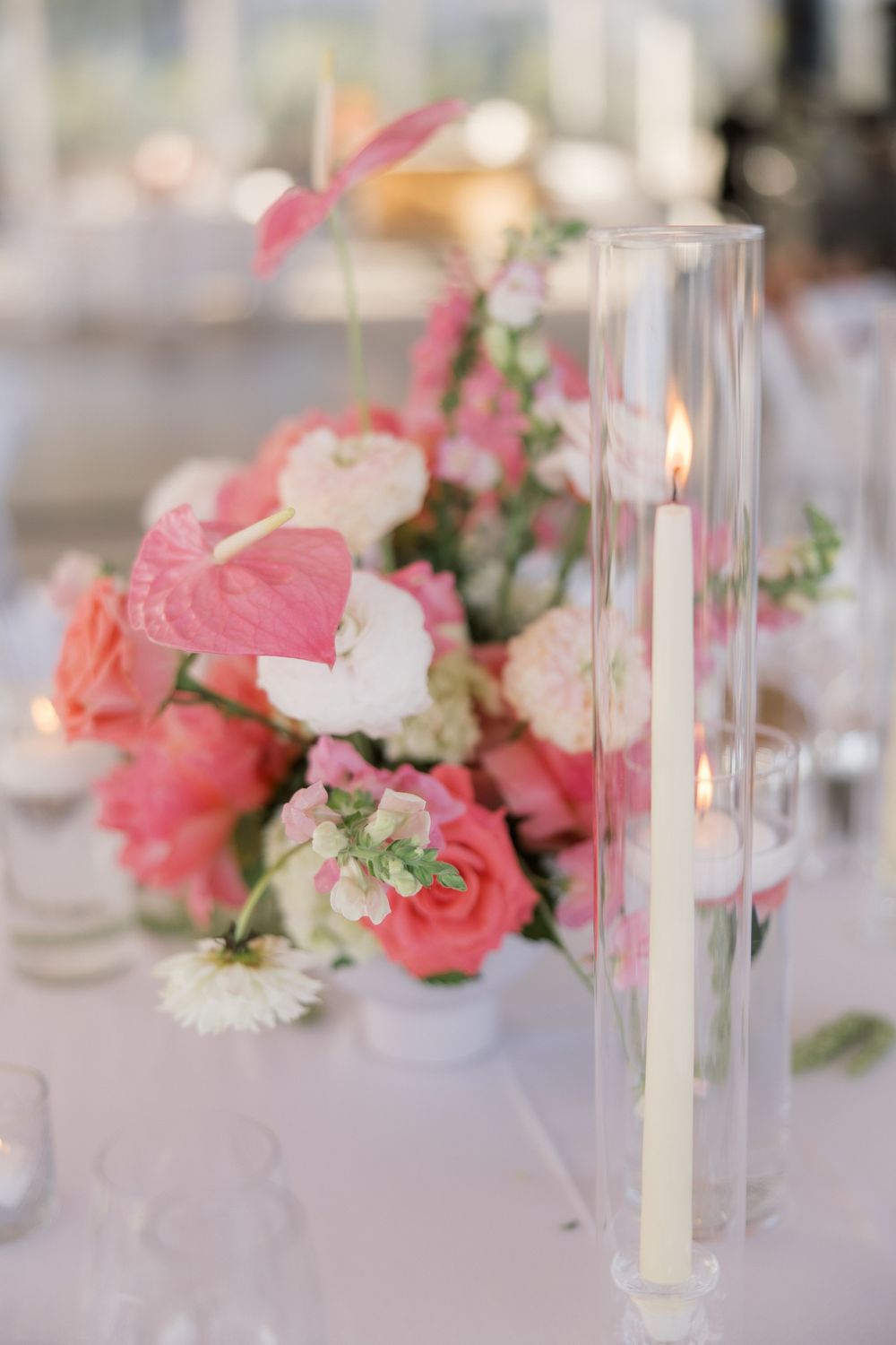Elegant wedding centerpiece with pink roses, anthuriums, and a tall glass candle holder with lit taper candle.