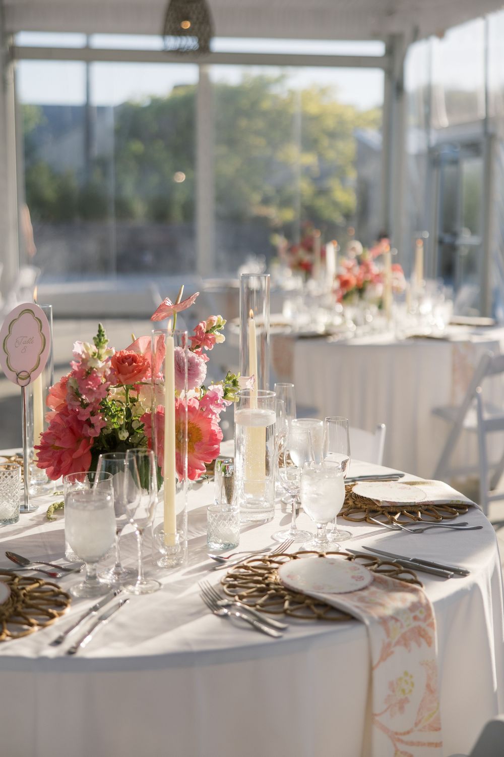 Elegant wedding reception table with pink floral centerpieces, taper candles, gold chargers, and crystal glassware in a bright venue.