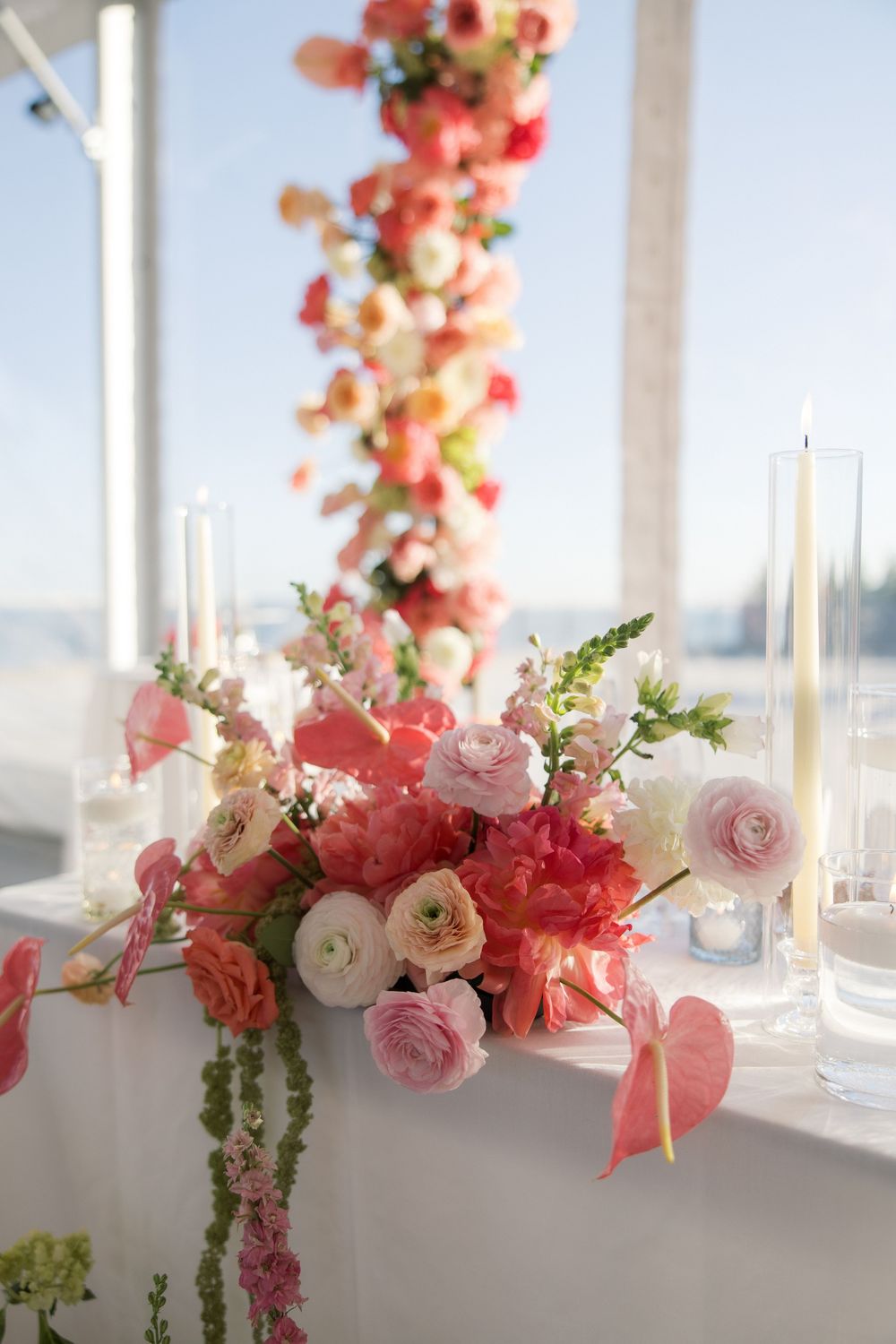 Elegant wedding table with pink and coral floral centerpiece, white candles, and cascading flower garland backdrop.