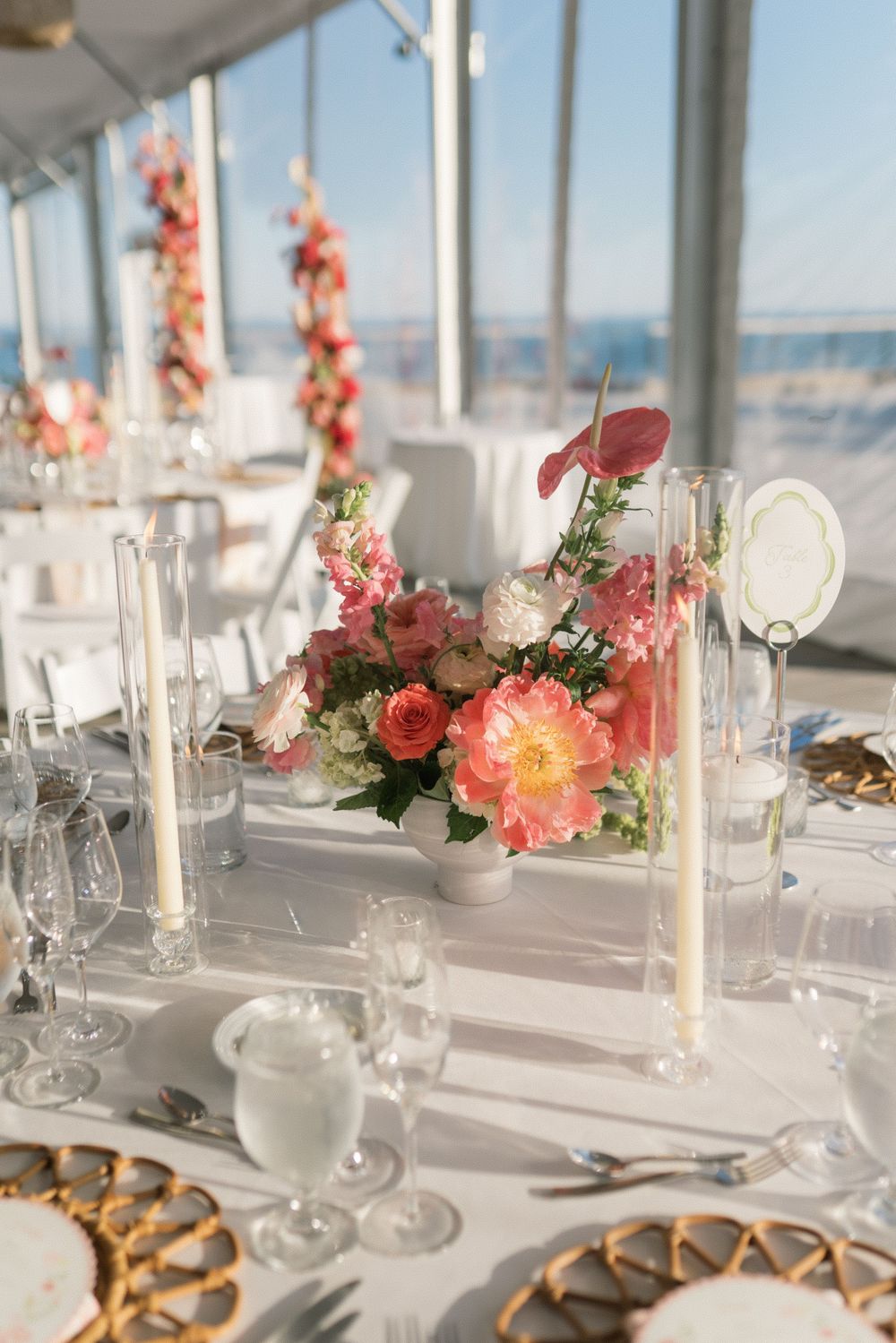 Elegant wedding reception table with pink floral centerpiece, tall candles, crystal glassware, and woven charger plates in a bright venue.
