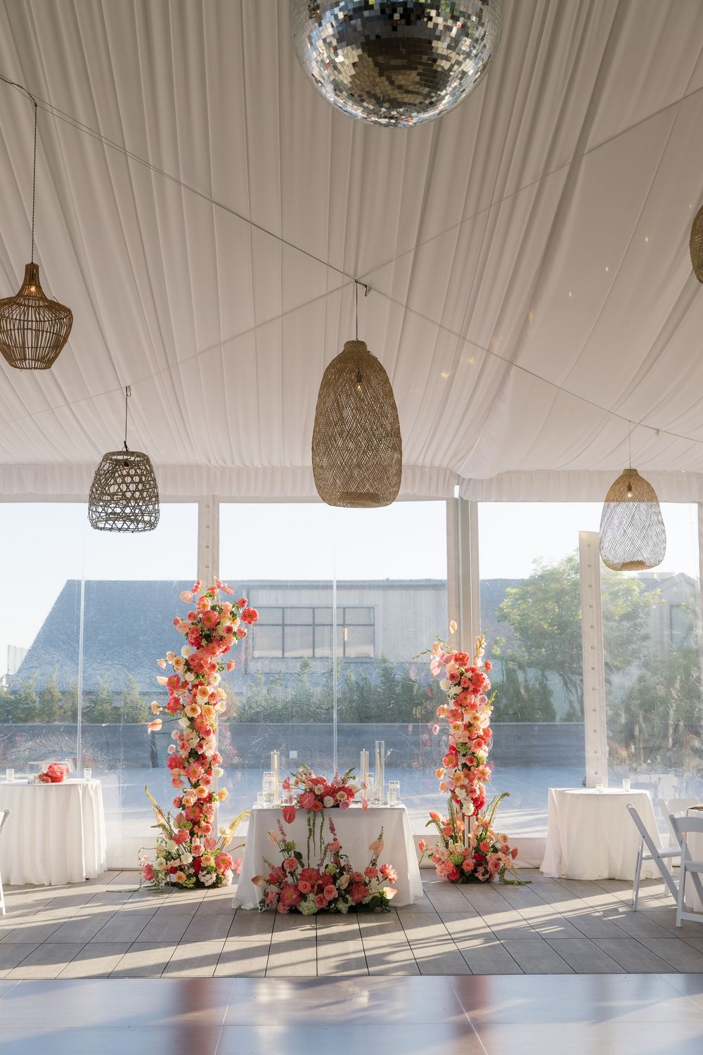 Elegant wedding reception tent with floral columns, disco ball, wicker pendant lights, and white draped ceiling.