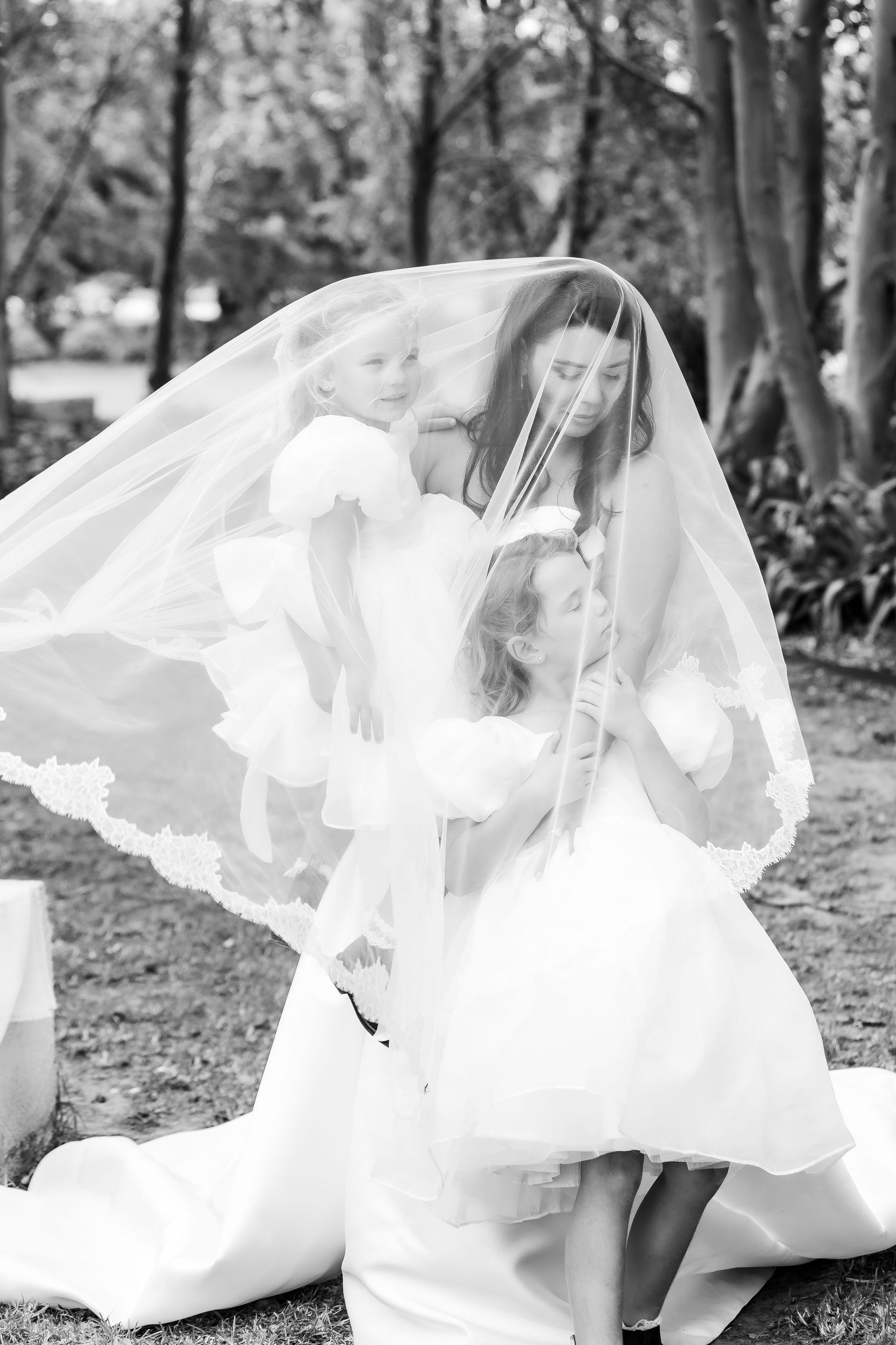 Black and white photo of a bride in a wedding veil with two young flower girls in a park setting.