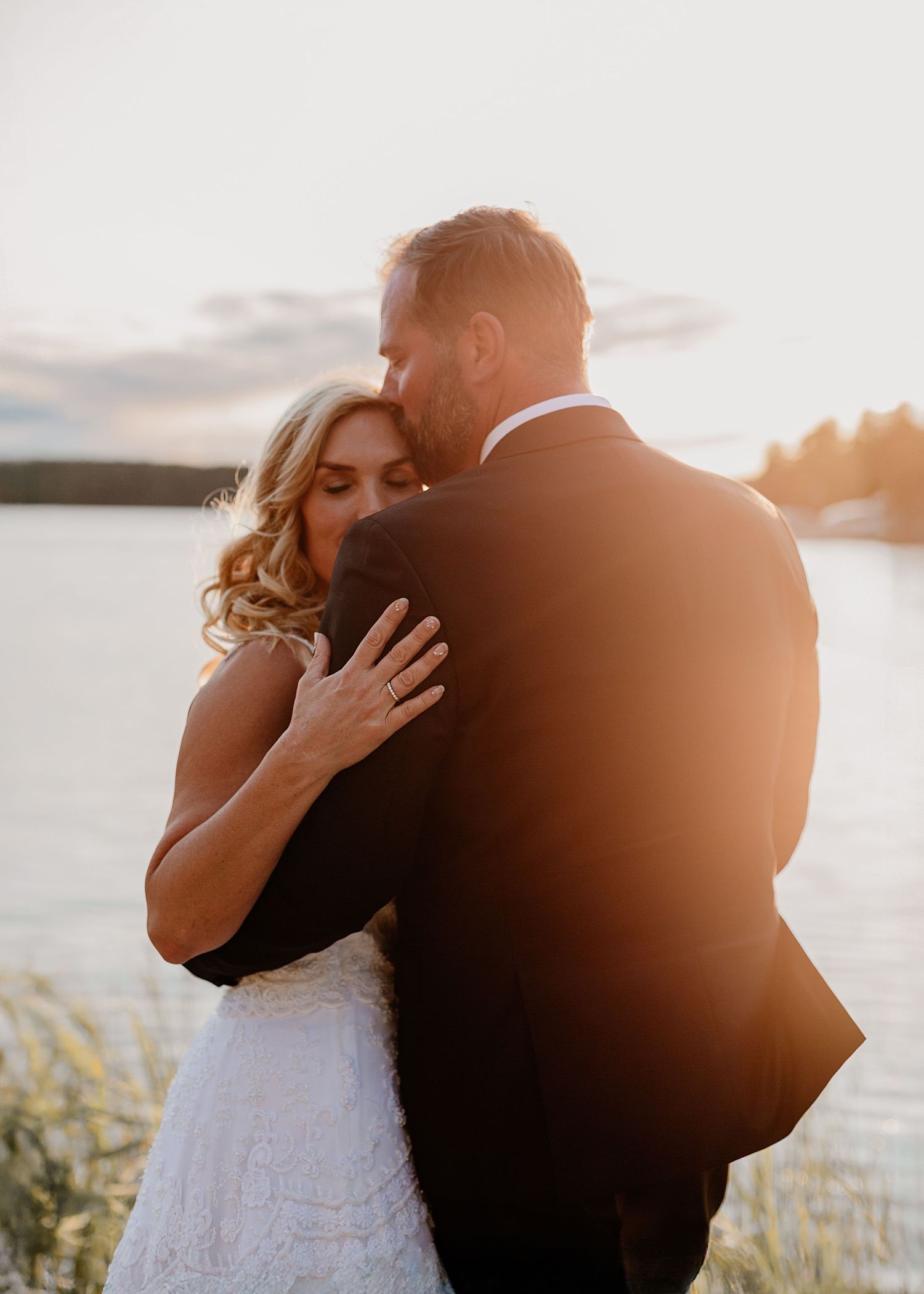 A couple shares a romantic embrace at sunset by a lake, the bride in white lace and groom in a black suit.