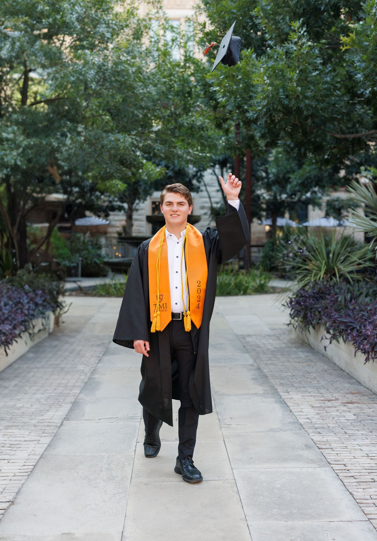 Smiling male graduate in black gown and orange stole holds cap toward camera outdoors on campus.
