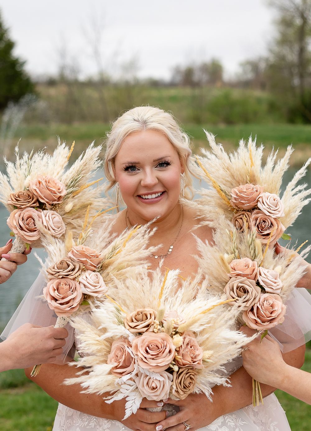 Smiling bride holding large boho bouquets of pampas grass and blush roses at outdoor wedding.
