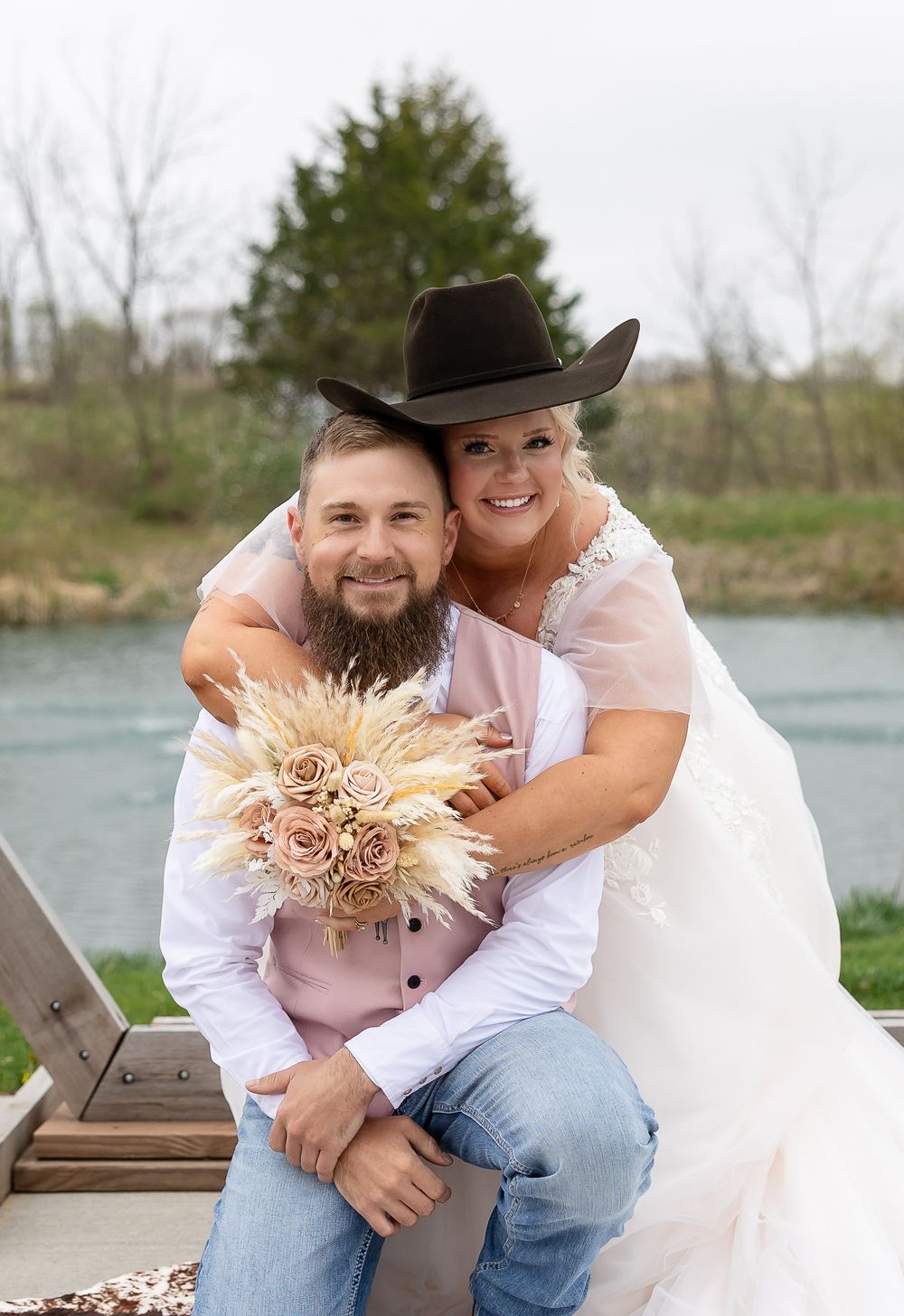 Smiling bride in white gown hugs bearded groom in cowboy hat holding blush bouquet at rustic wedding.
