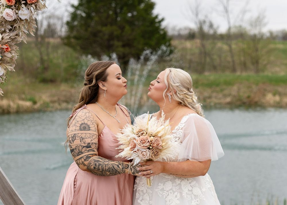 Two women in wedding attire share a tender moment near a pond, one in a white gown holding a bouquet.