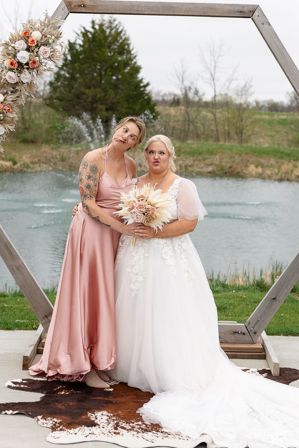 Bride in white lace gown and bridesmaid in pink satin dress pose under hexagonal wooden arch by a pond.