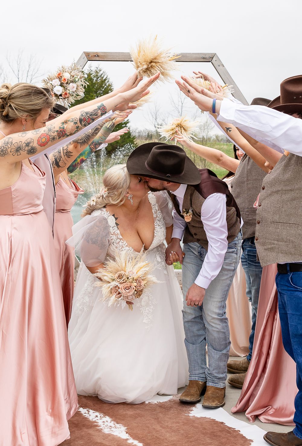 Bride and groom kiss under an arch as wedding party raises pampas grass in a rustic country-themed outdoor ceremony.