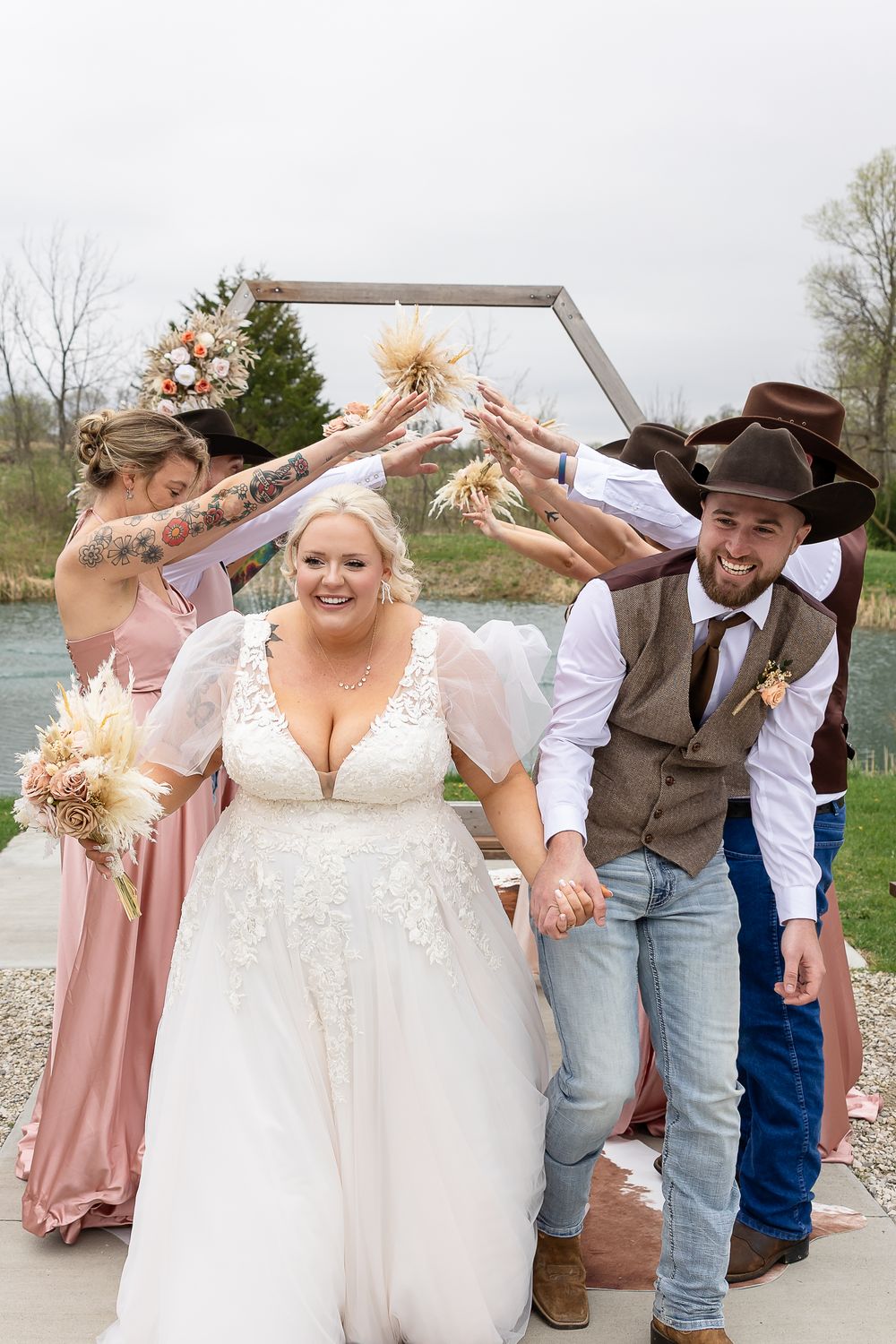 Smiling bride and groom walk under an arch of bouquets with bridesmaids at a rustic outdoor wedding.
