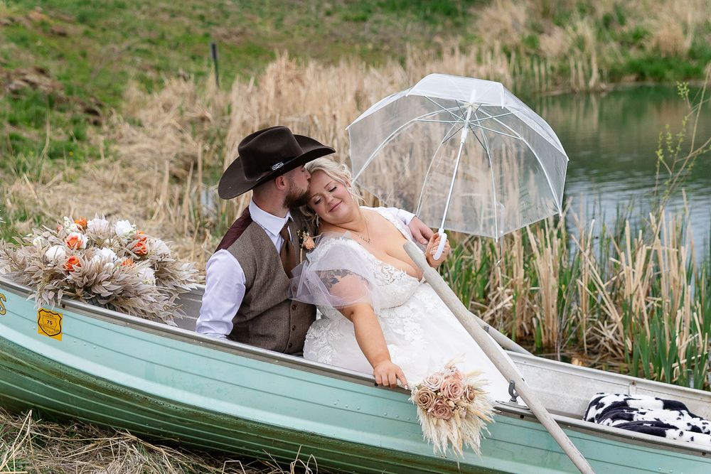 Bride and groom share a romantic moment in a canoe, holding a clear umbrella and bouquet near a peaceful pond.