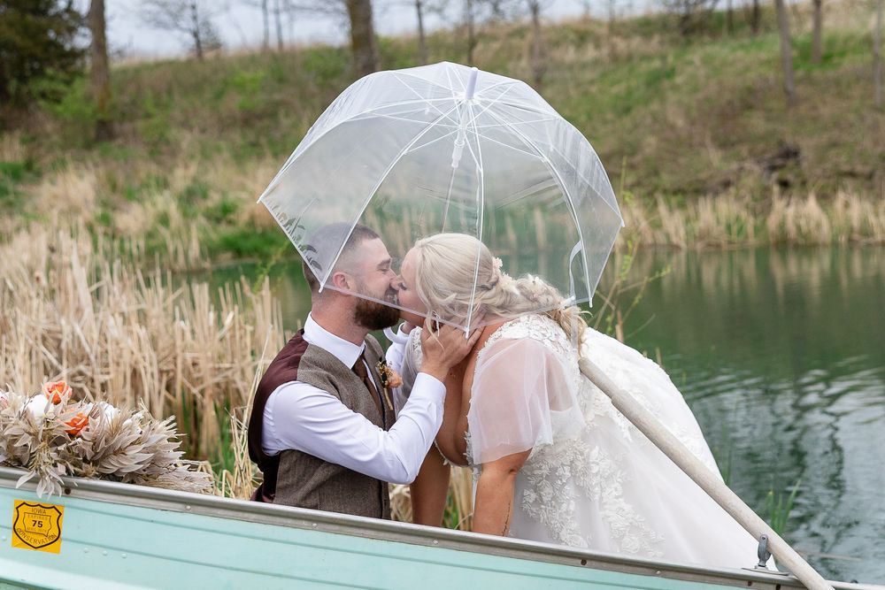 Bride and groom share a romantic kiss under a clear umbrella in a boat on a serene lake on their wedding day.