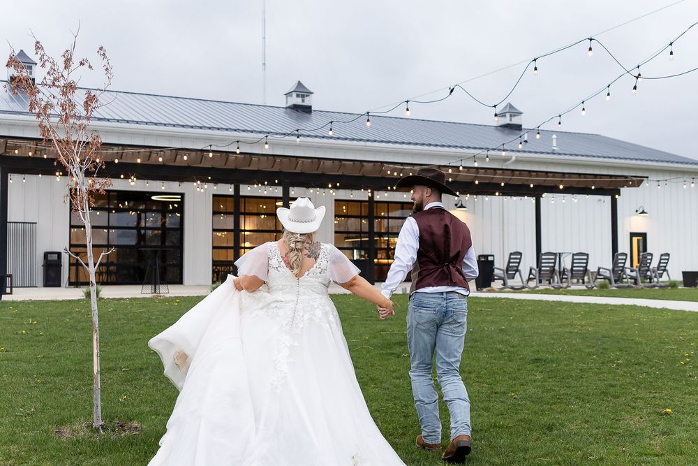 Bride in white gown and cowboy hat dancing with groom outside rustic barn wedding venue with string lights.